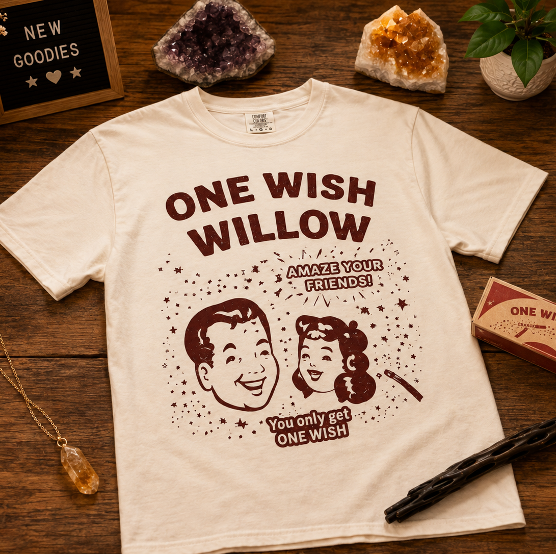 An off-white graphic t-shirt featuring a retro cartoon illustration of a man and woman with the text 'ONE WISH WILLOW' and 'AMAZE YOUR FRIENDS! YOU ONLY GET ONE WISH!' printed in dark brown. The t-shirt is displayed in a styled flat-lay arrangement on a wooden table surrounded by crystals, a candle, a necklace, and a vintage-style box.