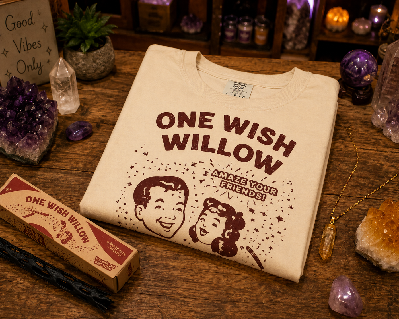 A cream-colored t-shirt with a retro-style brown graphic reading 'ONE WISH WILLOW' and 'ANZIE YOUR FRIENDS!' is displayed on a wooden table surrounded by crystals, a vintage-style box, and mystical props.